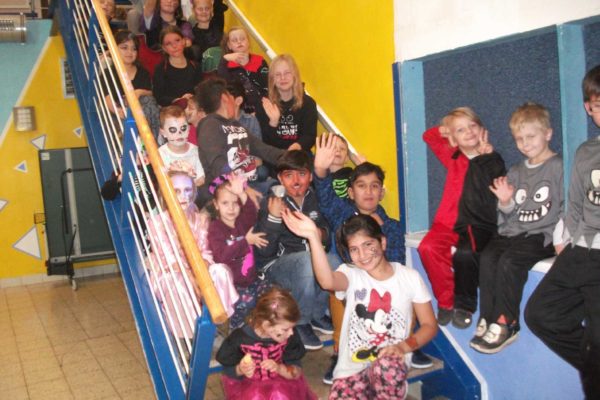 Viele Kinder sitzen gemeinsam auf einer Treppe und winken zum Teil. Sie sind zu Halloween verkleidet.